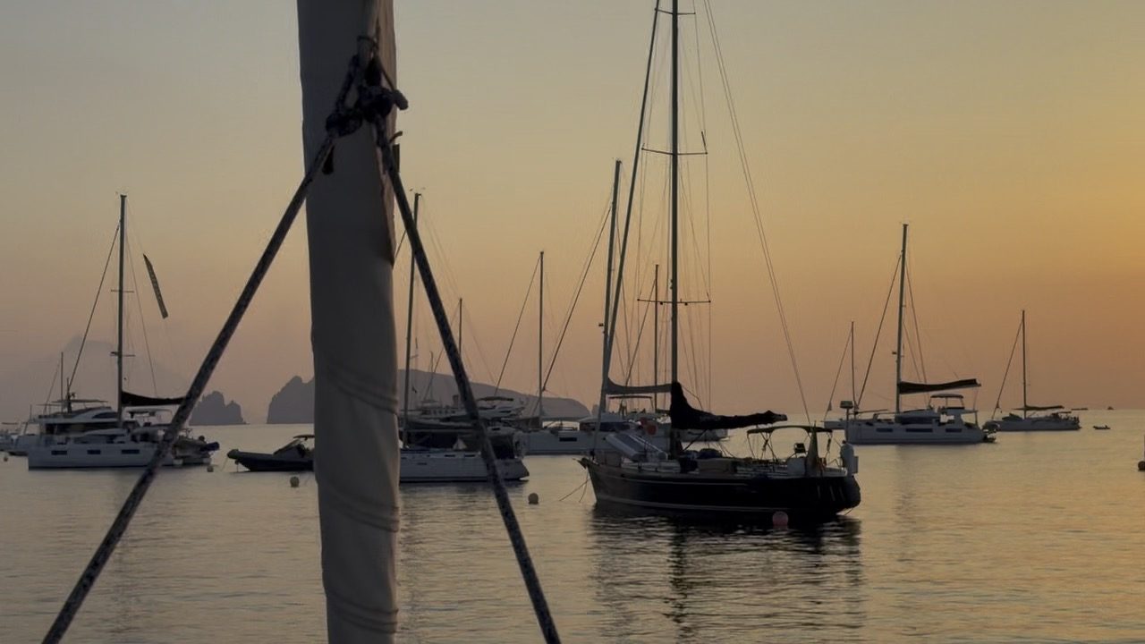 Yachts lying at anchor in flat water at sunset, an island headland on the horizon.